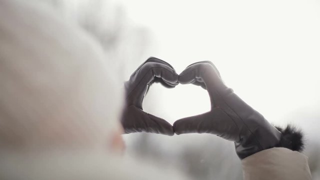 Women in beige down coat and hat making the symbol of love, heart with her hands in brown gloves against rising morning sun in snowy central park, slow motion. Back view