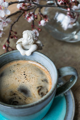 View of a white angel figurine hanging on a vintage cup of coffee on a rustic, old wooden table and blooming tree branches in a vase. Hello spring or Happy Easter background or concept. Toned image