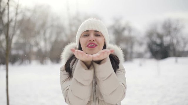 Brunette hair young women portrait with pink lips in beige down coat and hat sends an air kiss in snowy central park, snowfall. Slow motion.