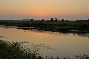 Sonnenuntergang an der Havel, Sonner f&auml;rbt den Himmel orangerot, Gr&auml;ser vom Ufer im Vordergrund. Vorherschende Farbe orangerot.