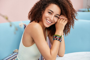 Sideways shot of pleased dark skinned young female has curly hair, broad smile, dressed in casual clothing, wears bracelet, sits on blue comfortable sofa in restaurant against cozy interior.