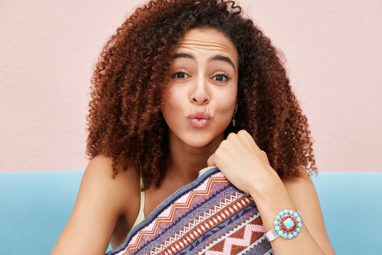 Candid shot of funny Afro American young woman makes grimce, rounds lips, has bushy curly hair, embraces cushion, spends free time in company of friends, have fun together, pose on sofa indoor