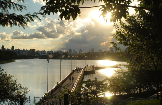 Represa municipal de S&atilde;o Jos&eacute; do Rio Preto ao p&ocirc;r do sol, com reflexos dourados na &aacute;gua, silhuetas de &aacute;rvores, passarela com pessoas e c&eacute;u parcialmente azul com nuvens