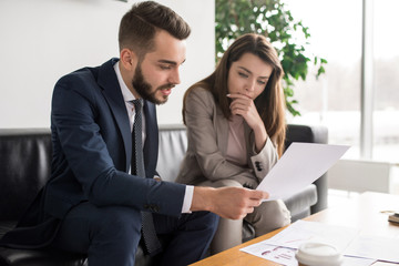 Portrait of two modern business people discussing documents in office, focus on pretty young woman reading contract