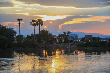 Abends am Mekong, Sonnenuntergang mit großer Farbpalette, Flussufer horizontal im Bild, Palmen am linken Uferrand, Hütten am rechten Rand, Farben der untergehenden Sonne spiegeln sich im Wasser
