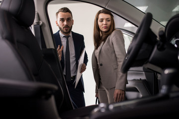 Portrait of handsome car salesman showing luxury car to woman in dealership showroom, shot from inside car