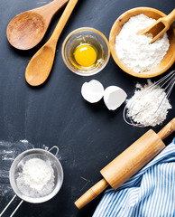 Baking ingredients. Bowl, eggs, flour, eggbeater, rolling pin and eggshells on black chalkboard from above.