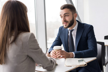 Portrait of two business people, man and woman, drinking coffee at table in cafe sitting at big window and talking