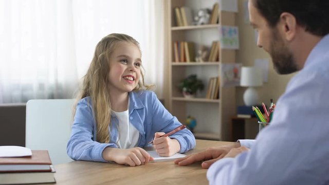 Smiling little daughter sitting at table and doing homework with father, care