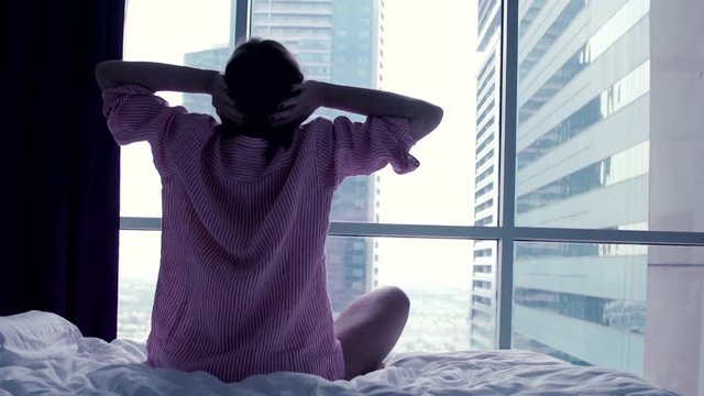 Young woman stretching her arms sitting on bed close to the window

