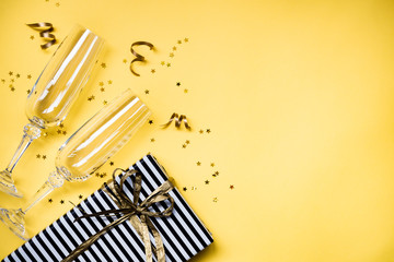 Celebration background - top view of two chrystal champagne glasses, a gift box wrapped in black and white striped paper, ribbons and star shaped golden confetti over yellow background. Copy space.