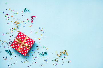 Top view of a red dotted gift box, scattered glittering star shaped confetti and colorful ribbons over blue background. Celebration concept. Copy space.