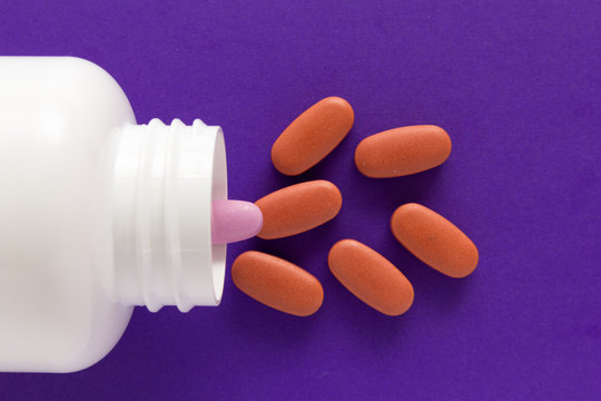 All tablets are red except one: pink. Capsules spilling out of white bottle. Purple background.