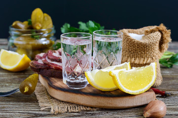 Two shot glasses of vodka with lemon slice, pickled cucumbers and rye bread with salted bacon on the dark background. Traditional strong drink and appetizer. Close-up.