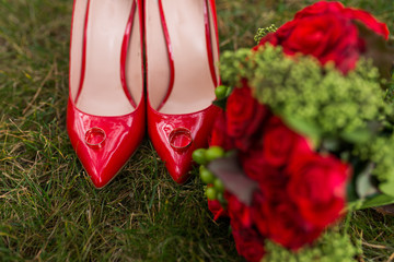 Two golden wedding rings lie on red fashion female shoes on green grass. Wedding details