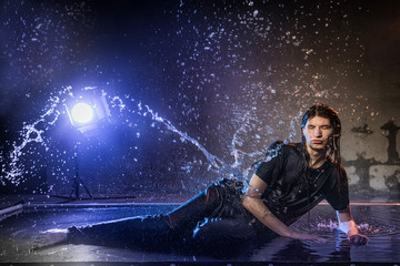 Attractive young man in black wet clothes under the rain and splash of water, studio photo