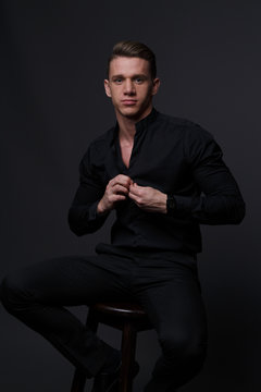 A white man sits on a dark chair in a black shirt and black mills, on a gray background