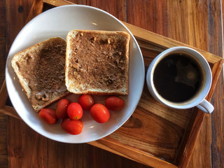 Whole wheat bread with peanut butter and tomatoes in white dish with Black coffee cup on wooden background.Top view and natural light.