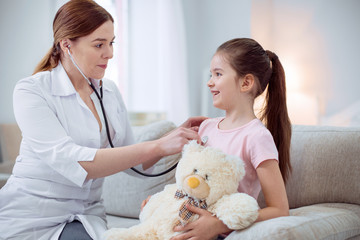 Health problem. Merry nice girl sitting on couch and hugging plush bear while female doctor using stethoscope
