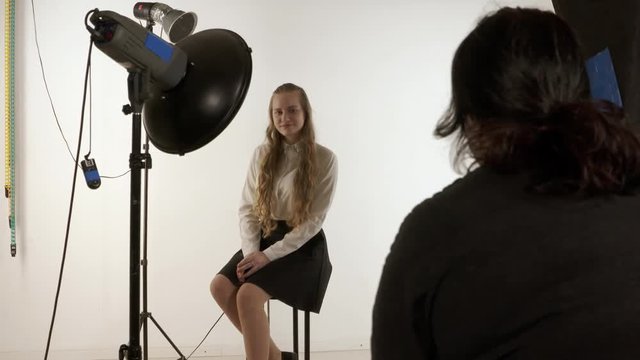 Photographer works with blonde young model in photo studio. Woman photographing teen girl in white shirt. Backstage shot of photoshoot in a studio.