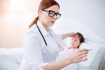 Fototapeta premium Terrible fever. Worried upset female doctor estimating body temperature and posing on blurred background where girl lying
