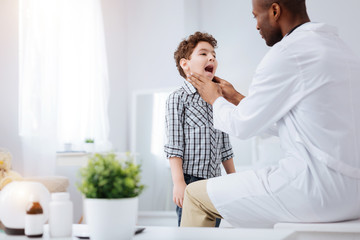 Little patient. Appealing charming boy catching cold while showing throat to doctor and opening mouth