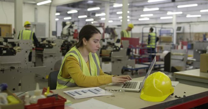 A female manager on a laptop in a busy production factory.