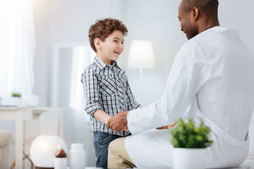 Struggle with cold. Skillful male doctor sitting while talking to jovial boy who laughing