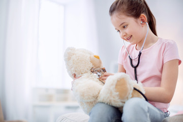 Future doctor. Vigorous adorable kind girl hearing plush bear while sitting on light background and staring down