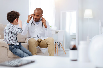 Symptoms and causes. Charming nice male doctor and boy sitting on sofa while communicating and pointing at ears