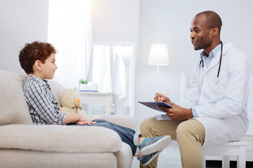 Your complains. Gay afro American male doctor listening to boy while holding clipboard and wearing uniform