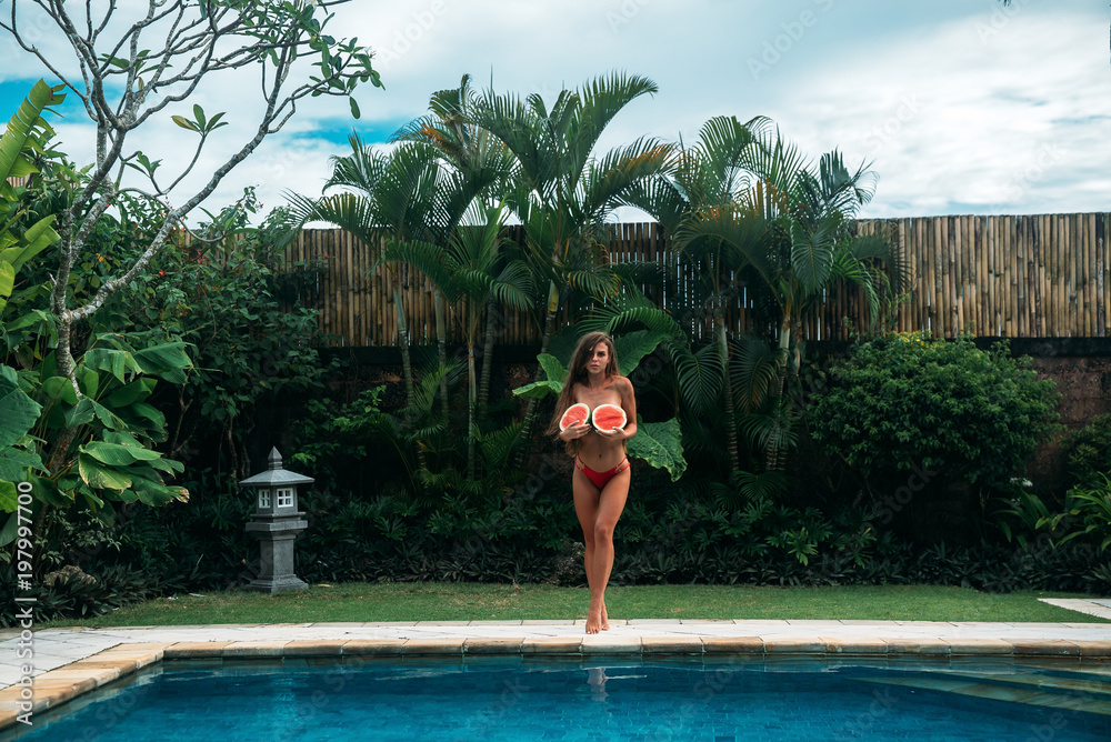 Wall mural cute tanned girl in a red bikini with curly long hair against a background of large green tropical l