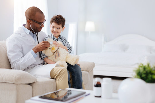 Plush toy. Male doctor sitting with cheerful jolly boy while examining plush bear and communicating