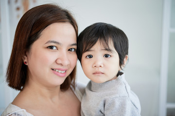 Head and shoulders portrait of beautiful Asian woman looking at camera with wide smile while holding her cute little son on hands, blurred background