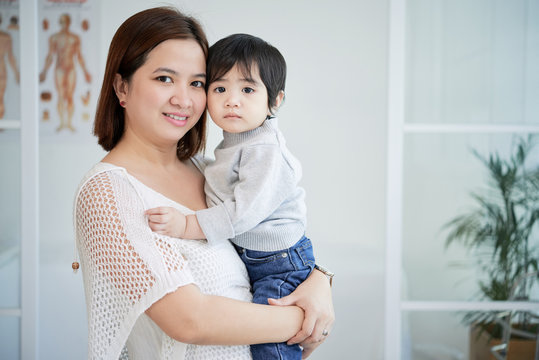 Group portrait of pretty Asian woman holding her cute little son on hands and posing for photography while standing at pediatrician office