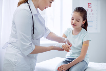 Broken arm. Charming smart interested girl broking arm and female doctor putting plaster while standing