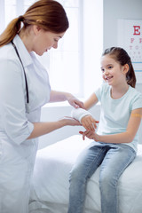 Fototapeta premium Too active girl. Happy joyful girl broking arm and female doctor applying plaster while staying