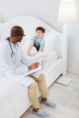 Obraz premium Symptoms description. Top view of nice boy describing his symptoms while afro american male doctor sitting on bed and using clipboard