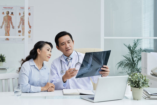 Handsome middle-aged surgeon sitting at desk and showing X-ray image to senior patient while discussing possible treatment, interior of modern office on background
