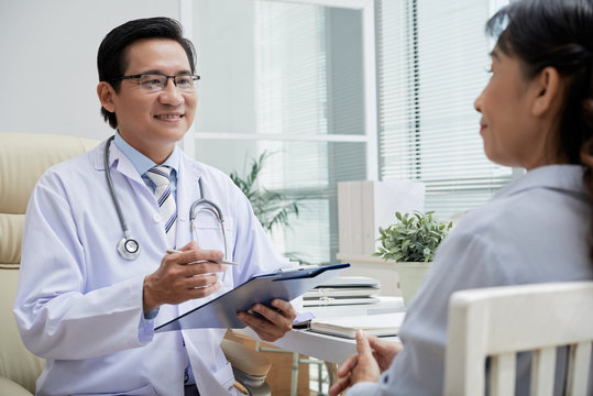 Friendly middle-aged physician wearing white coat sitting opposite senior patient and giving recommendations while having appointment at modern office