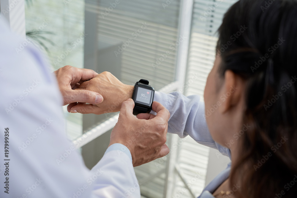 Wall mural Over shoulder view of friendly therapist showing his senior patient how to check heart rate with help of smartwatch, close-up shot