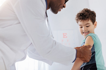 Just scratch. Low angle of male doctor placing patch while pretty little boy gazing at hand and sitting