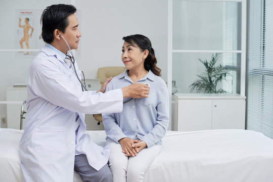 Profile view of confident middle-aged therapist wearing white coat using stethoscope while examining senior patient, interior of modern ward on background