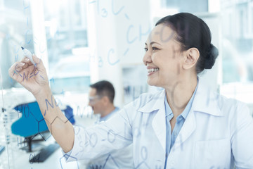 Interesting research. Beautiful cheerful dark-haired experienced writing formulas and wearing a uniform while working in the lab with her colleague