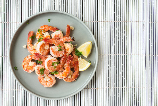 Top view of cooked delicate shrimps with pieces of lemon and cut fresh greens on grey platter. Well-served, full of vitamins and diet dish. Copy space in right side