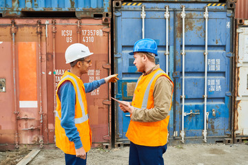 Side view portrait of two dock workers wearing hardhats talking to each other standing against cargo containers