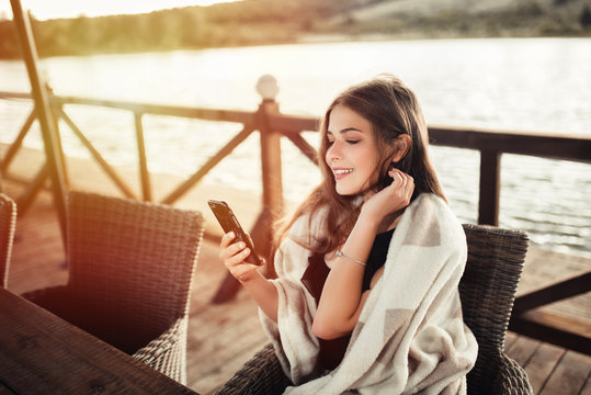 Cheerful relaxed woman uses smart phone for chat with friends, sits in modern coffee shop or terrace cafe. Pretty female reads good news on internet website, enjoys summer rest.