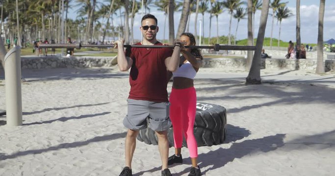 Content sportive woman in activewear supporting man while doing squats with barbell in bright sunlight on sandy beach. 