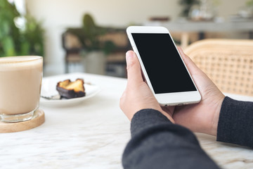 Mockup image of hands holding white mobile phone with blank black screen with a cup of coffee and snack on table in cafe