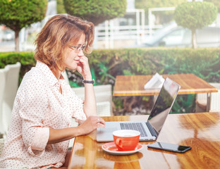 Young attractive girl in glasses with a laptop and a cup of coffee in a street cafe, online shopping, payments, remote work